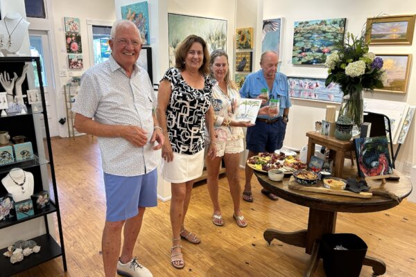 Four adults smile and stand near a table with food and drinks in an art gallery filled with paintings, jewelry, and flowers. The setting appears casual and cheerful.