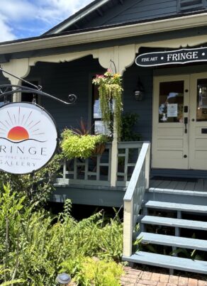 A blue house with a porch hosts the Fringe Art Gallery. A large round sign with a sun logo hangs by the steps, surrounded by greenery, flowers, and hanging plants. The sky is partly cloudy.