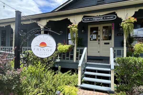 A blue house with a porch hosts the Fringe Art Gallery. A large round sign with a sun logo hangs by the steps, surrounded by greenery, flowers, and hanging plants. The sky is partly cloudy.