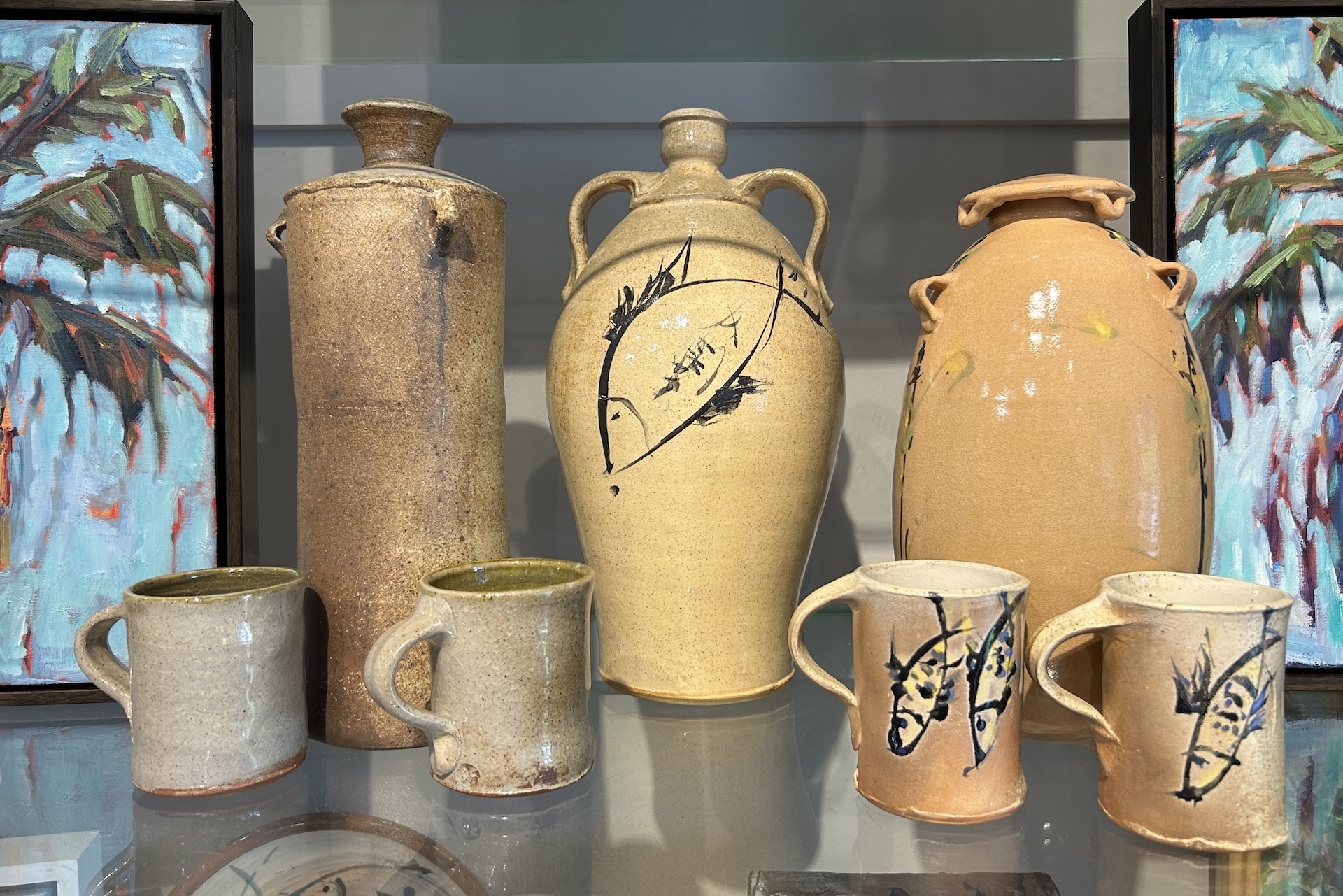 Five pottery pieces, including two tall vases and three mugs, are displayed in front of two colorful paintings with palm tree designs. Two mugs and one vase feature fish illustrations on their surfaces.