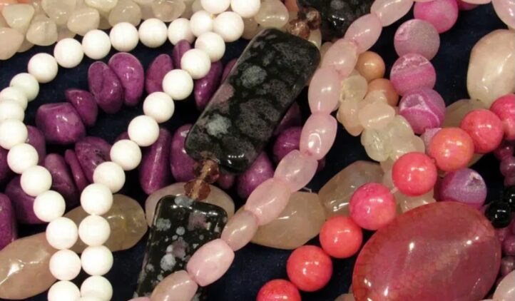 A close-up of assorted colorful gemstone and bead necklaces, including white, pink, purple, black, and peach beads in various shapes and sizes, all overlapping on a dark background.