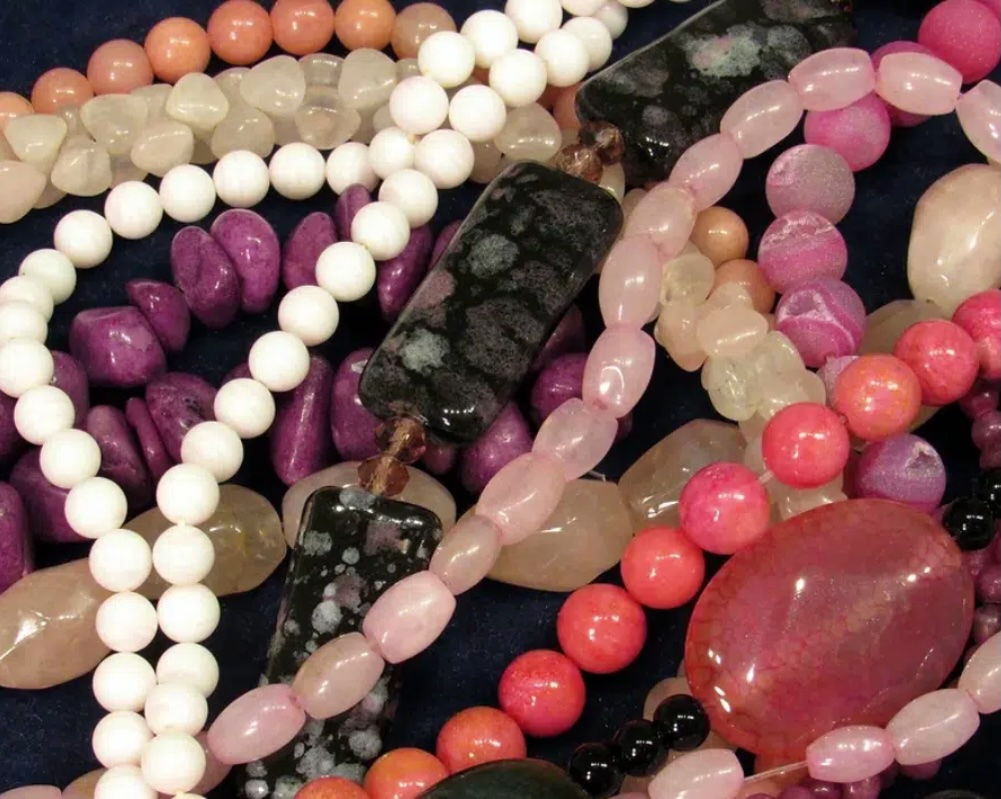 A close-up of assorted colorful gemstone and bead necklaces, including white, pink, purple, black, and peach beads in various shapes and sizes, all overlapping on a dark background.