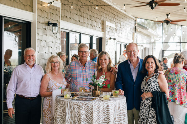 Six adults, three men and three women, stand smiling around a cocktail table with drinks, floral centerpiece, and magazines on an outdoor patio decorated with string lights. People and greenery are visible in the background.