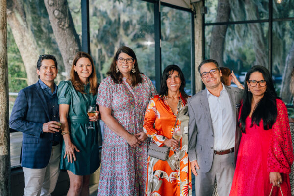 Six people dressed in colorful, semi-formal attire stand together indoors, smiling at the camera. Some hold drinks, and large windows behind them show trees and greenery outside.