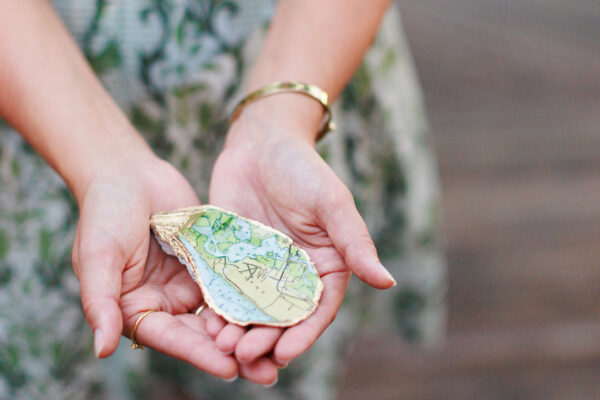 A person wearing a gold bracelet and rings holds an oyster shell painted with a colorful map. The background is out of focus, highlighting the shell and the persons hands.
