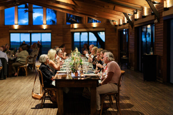 A group of people sit at a long wooden table in a warmly lit, rustic venue with large windows, enjoying dinner and conversation as evening light shines outside.