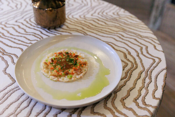 A white plate with a circular rice dish topped with diced vegetables and herbs, surrounded by a ring of green sauce. The plate sits on a table with a wavy, striped pattern. A small brown vase is in the background.
