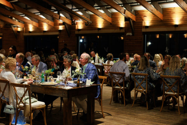 A large group of people sit at wooden tables in a warmly lit indoor venue, engaged in conversation and dining. The room features wooden beams, plants, and decorated tables.