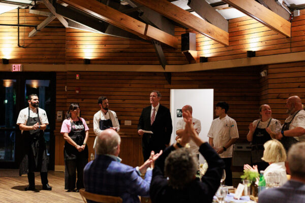 A group of chefs and a man in a suit stand at the front of a wooden event hall, while seated guests clap and watch. Warm lighting highlights the scene.