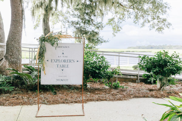A sign reading “At the Explorer’s Table” is displayed on a metal stand, decorated with greenery and tan foliage. It stands outdoors by trees and plants, overlooking a body of water under a cloudy sky.