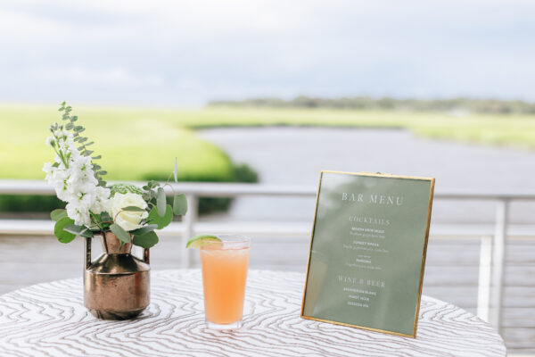 A table with a copper vase of white flowers, a pink cocktail with a lime wedge, and a framed bar menu sits outdoors by a river and green marshland under a cloudy sky.