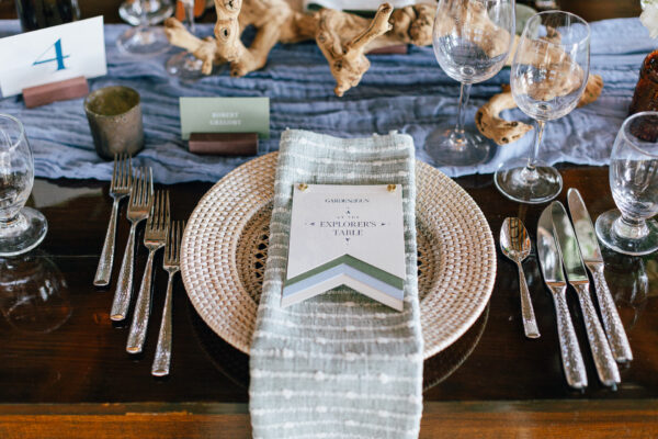 A place setting on a wooden table with a woven charger, gray striped napkin, silver cutlery, glassware, and a card reading Explorers Table. Driftwood and blue cloth runner decorate the table.