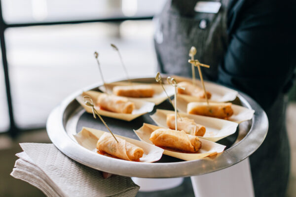 A server holding a round tray with small plates, each containing a crispy spring roll secured with a bamboo pick, ready to be served as appetizers at an event. A stack of napkins rests on the trays edge.