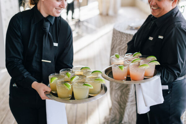 Two servers in black uniforms hold trays of cocktails garnished with lime wedges. The servers are smiling, and each holds a tray with several drinks, set in a bright, indoor setting.
