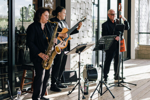 Three musicians perform on a wooden patio: a woman playing the saxophone, a man playing guitar, and another man playing an upright electric bass. All wear black suits and stand near music stands and audio equipment.