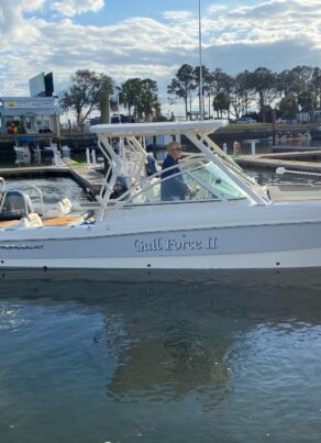 A white powerboat named Gulf Force II is on the water near a marina, with a person at the helm. Palm trees, docks, and other boats are visible in the background under a partly cloudy sky.