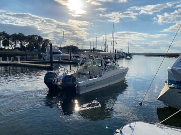 A white motorboat with two outboard engines is docked in a calm marina, surrounded by other boats and sailboats. The sun shines through scattered clouds, reflecting on the water. Trees and docks are visible in the background.