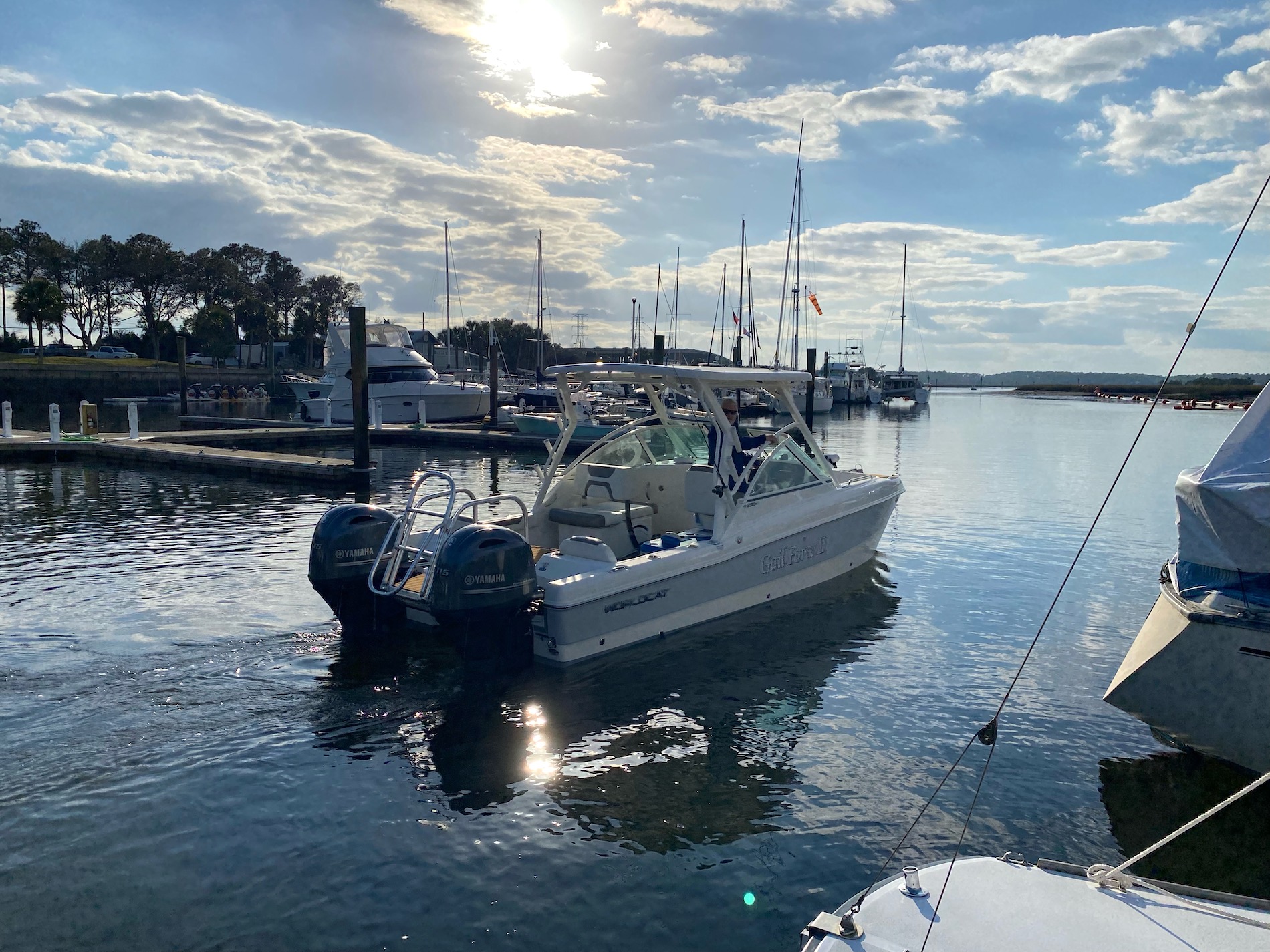 A white motorboat with two outboard engines is docked in a calm marina, surrounded by other boats and sailboats. The sun shines through scattered clouds, reflecting on the water. Trees and docks are visible in the background.