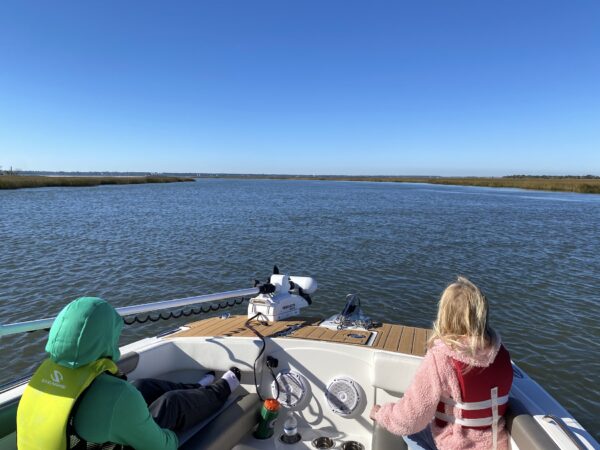 Two children wearing life jackets sit at the front of a boat on calm water, facing forward. The sky is clear and blue, and grassy banks line both sides of the waterway ahead.