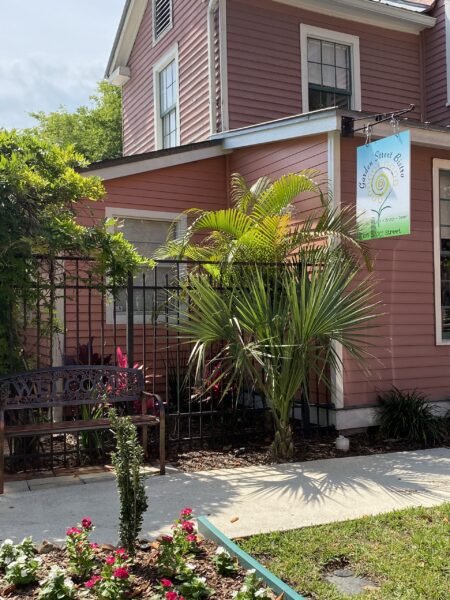 A pink wooden house with white trim features a black metal gate, a palm tree, a bench, and a blue sign reading “Linda Street Bistro.” Flowers and greenery decorate the yard under a sunny sky.