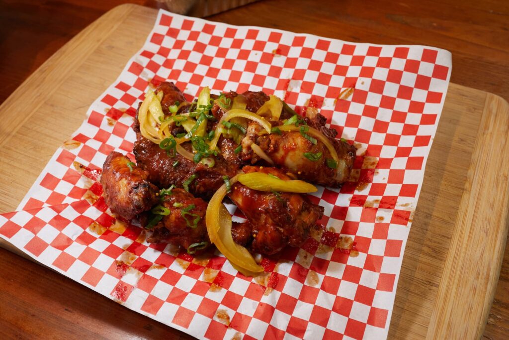 A serving of saucy chicken wings garnished with sliced green onions and yellow peppers, placed on red and white checkered paper atop a wooden cutting board.