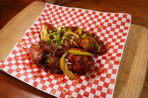 A serving of saucy chicken wings garnished with sliced green onions and yellow peppers, placed on red and white checkered paper atop a wooden cutting board.