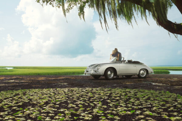 A couple sits in a classic white convertible parked under a large tree with hanging moss, overlooking a serene marshland with green grasses and blue sky in the background.