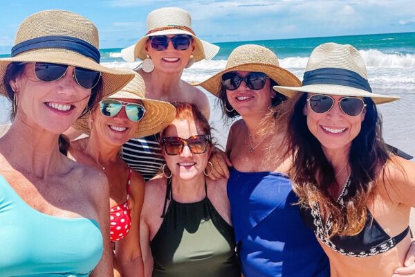 Six women in swimsuits and sun hats smile together for a group selfie on a sunny beach, with ocean waves and blue sky in the background.