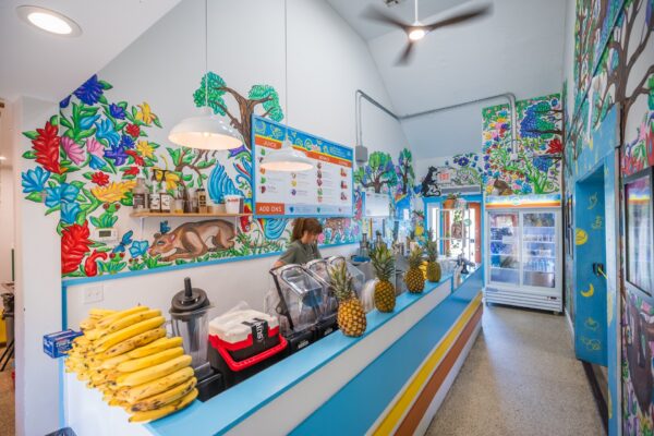 A colorful juice bar with vibrant tropical murals, a counter lined with bananas and pineapples, blenders, a menu board above, and a person preparing drinks near a refrigerator display in the background.