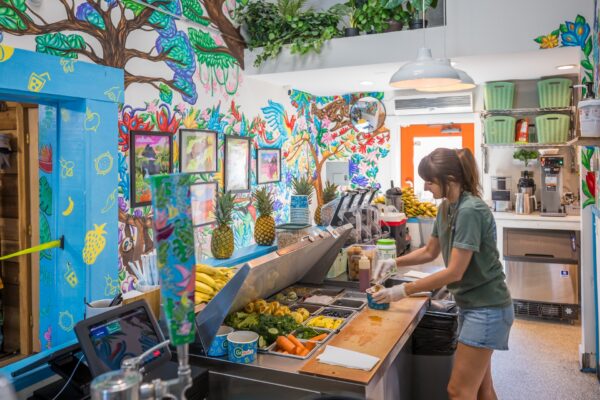 A woman prepares fresh fruit juice behind a counter in a colorful, tropical-themed juice bar decorated with vibrant murals, plants, and framed art. Bananas and pineapples are visible on the counter.