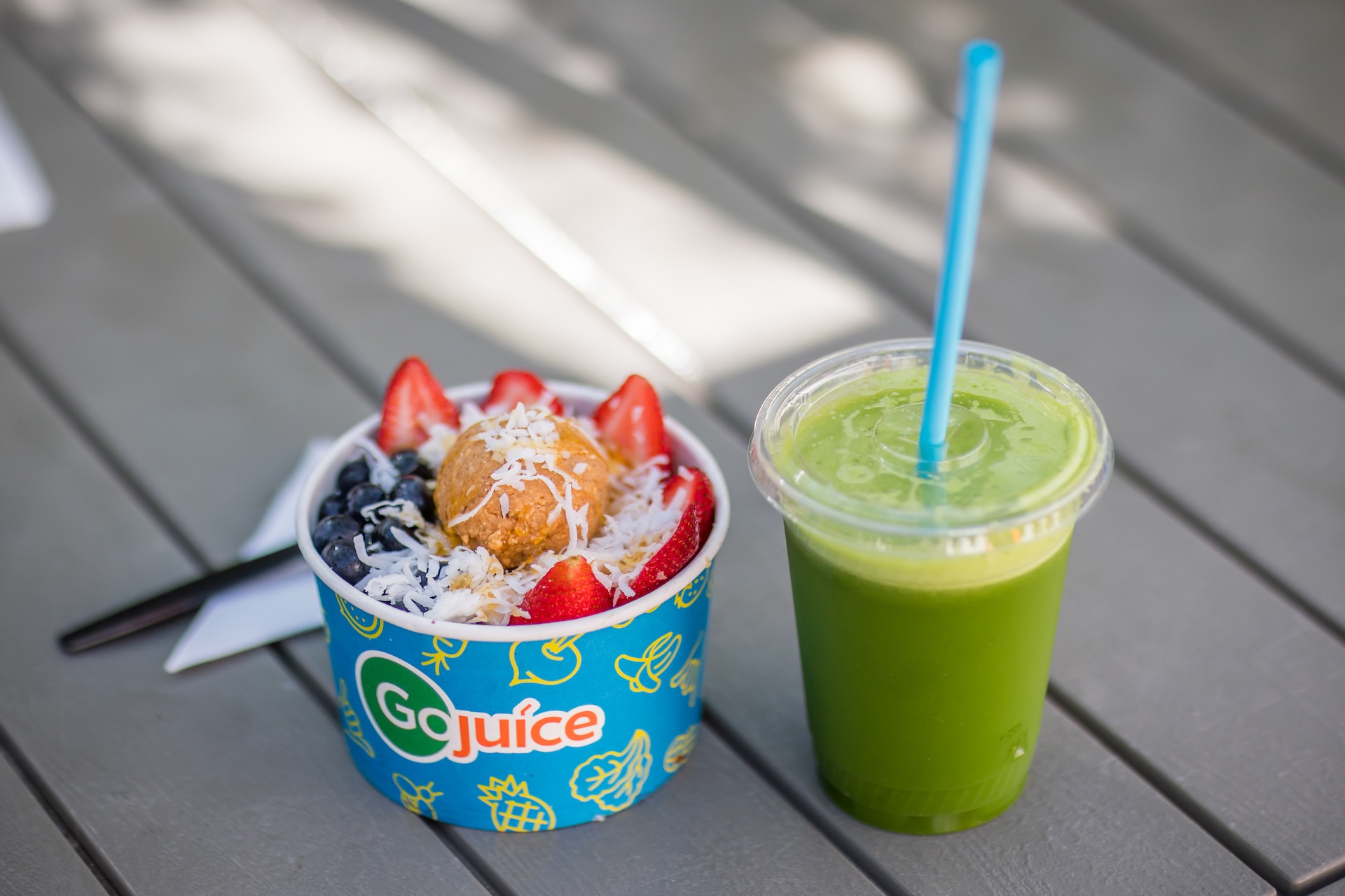 A bowl of acai topped with blueberries, strawberries, shredded coconut, and a round energy bite sits next to a cup of green juice with a blue straw on a gray wooden table.