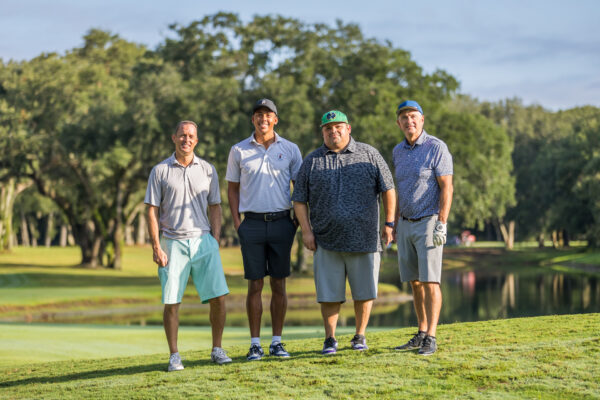 Four men stand side by side on a golf course near a pond, dressed in casual golf attire, with trees and grass visible in the background on a sunny day.