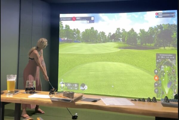 A woman stands indoors, poised to swing a golf club at a virtual golf simulator screen showing a green course. In the foreground are a glass of beer, a glass of wine, snacks, and papers on a wooden table.