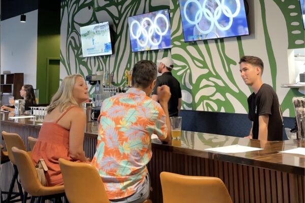 Two people sit at a bar talking to a bartender. Behind them are two TVs showing the Olympic rings, and the wall has a green abstract leaf design. The bartender listens attentively. Drinks are on the bar.