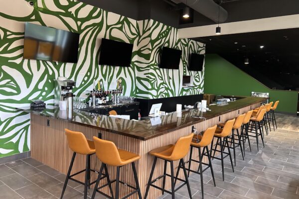 A modern bar with a glossy dark counter, orange barstools, and a tropical green and white leaf-patterned wall. Four TVs are mounted above the bar, and various drinks and glasses are arranged behind the counter.