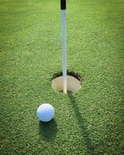 A close-up of a white golf ball resting on green grass right next to a golf hole with a flagstick in the center of the hole.