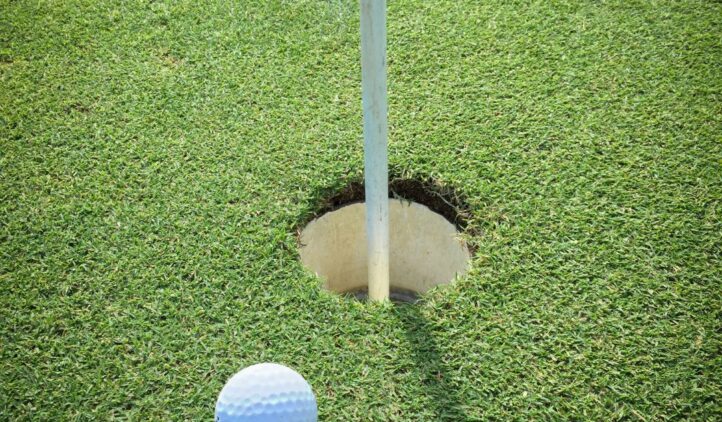 A close-up of a white golf ball resting on green grass right next to a golf hole with a flagstick in the center of the hole.