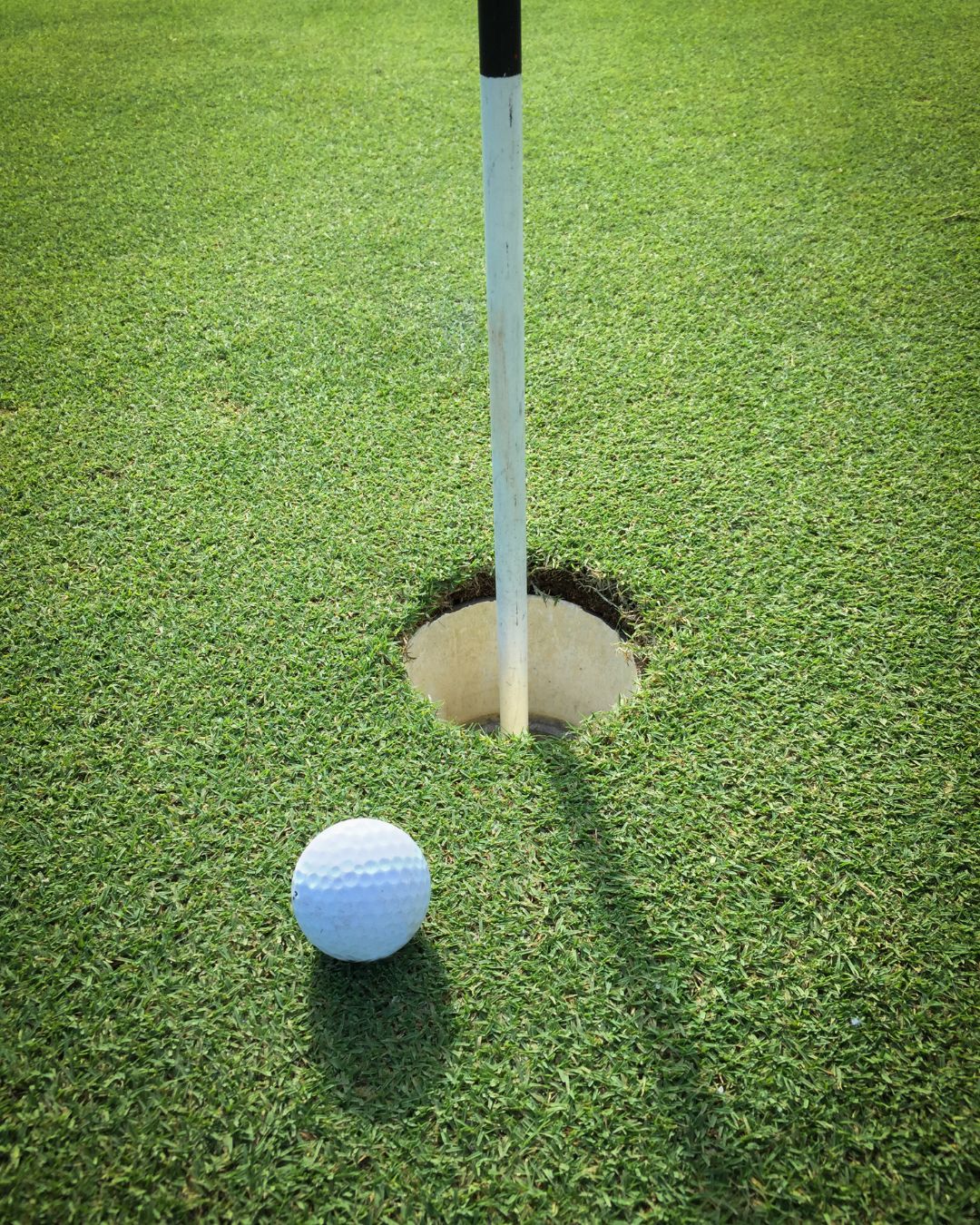 A close-up of a white golf ball resting on green grass right next to a golf hole with a flagstick in the center of the hole.