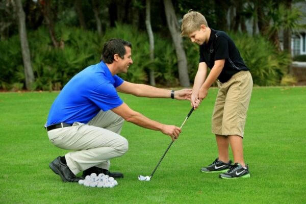 A man kneeling on grass helps a smiling boy position his hands on a golf club, with a pile of golf balls nearby and trees in the background.