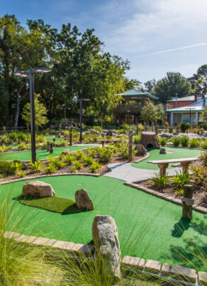 A landscaped outdoor mini golf course with green artificial turf, stone barriers, benches, pathways, and trees under a sunny sky.