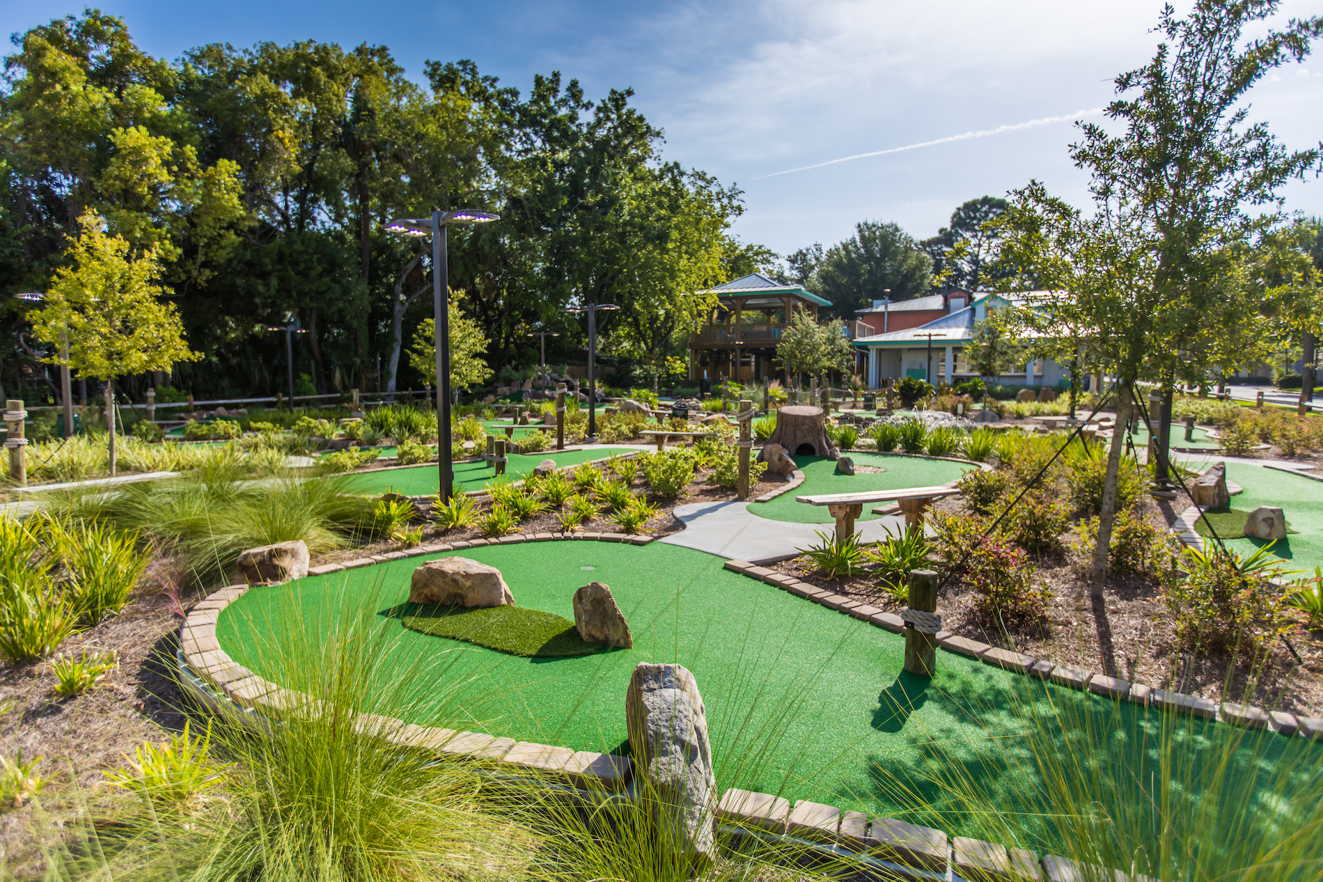 A landscaped outdoor mini golf course with green artificial turf, stone barriers, benches, pathways, and trees under a sunny sky.