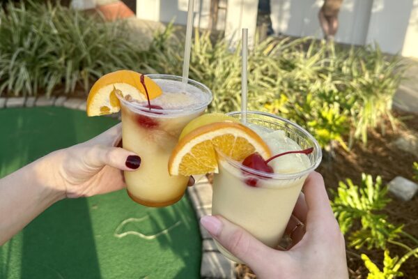 Two hands holding frozen cocktails garnished with orange slices and cherries, outside near greenery and a mini-golf course.