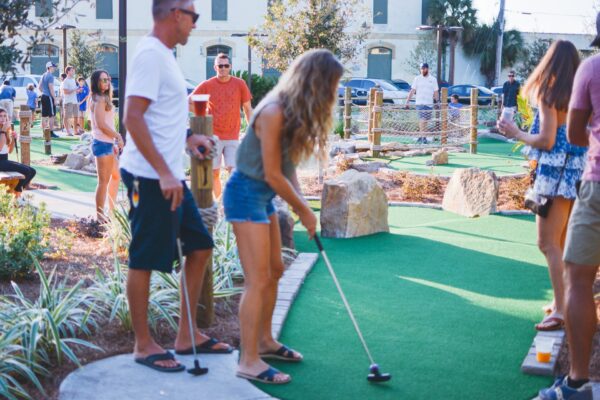 A group of people play mini-golf outdoors on a sunny day. In the foreground, a woman bends to hit a golf ball while others stand nearby, watching and holding putters. Green turf, plants, and rocks surround the course.