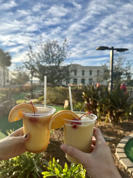 Two hands hold up tropical drinks garnished with orange slices and cherries under a bright blue sky, with greenery, trees, and a white building in the background.