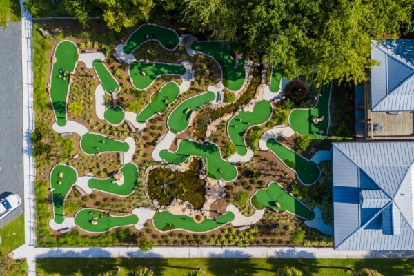 Aerial view of a mini golf course with winding green paths, sandy areas, landscaping, and a central pond, surrounded by trees and bordered by buildings and a parking lot.