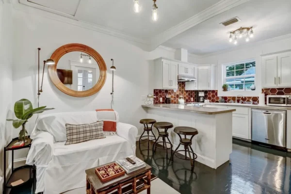 A cozy, modern kitchen and living space with white cabinets, a breakfast bar with three stools, a white sofa, decorative pillows, a round wall mirror, and hexagonal brown backsplash tiles.