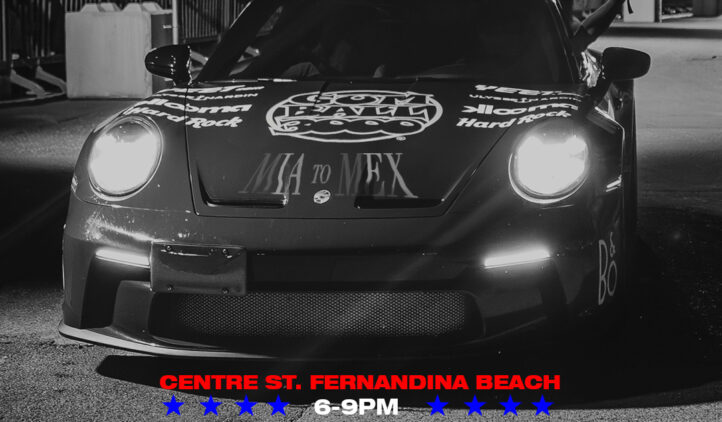 A black Porsche sports car at night on a street, surrounded by barriers and people. Text on the image reads: Gumball 3000 comes to Amelia Island, Centre St. Fernandina Beach, 05 June, 6–9 PM, 100 supercars & superstars.