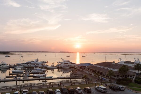 A marina at sunset with boats docked in calm water, a building on the right, parked cars in the foreground, and the sun reflecting on the water under a partly cloudy sky.