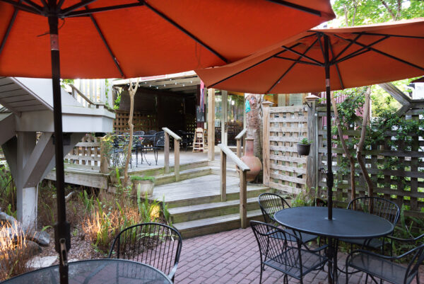 Outdoor patio with black metal tables and chairs under large orange umbrellas, surrounded by wooden fencing and greenery, with steps leading up to a covered area with more seating.