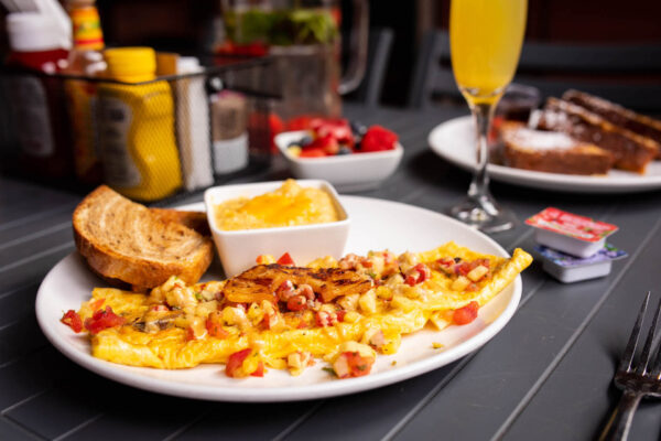 A plate with an omelet topped with diced vegetables, toast, a bowl of grits, and fresh fruit. In the background, there’s a drink, syrup packets, condiments, and a plate of French toast with powdered sugar.
